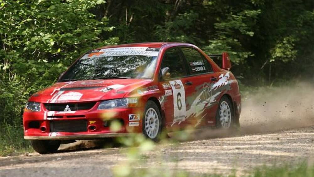 Automobiļu sacīkšu auto ar sporta numuru 6, brauc pa grants ceļu meža vidū.